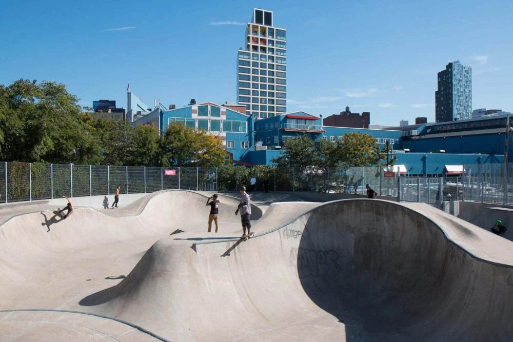 Skaters riding transitions at a large concrete skatepark in New York City with tall buildings in the background.