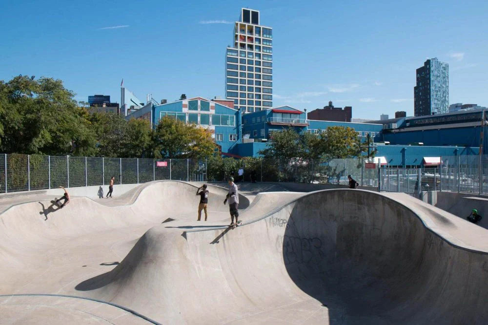 Skaters riding transitions at a large concrete skatepark in New York City with tall buildings in the background.