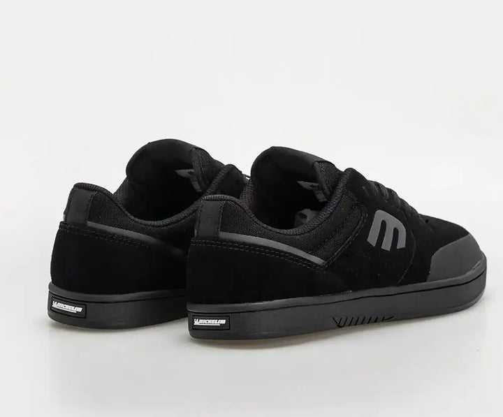 A pair of black low-top sneakers shown from a three-quarters rear angle on a light gray studio background. The shoes feature a mix of suede and textile materials, black laces, a black sole, and a gray logo on the side.
