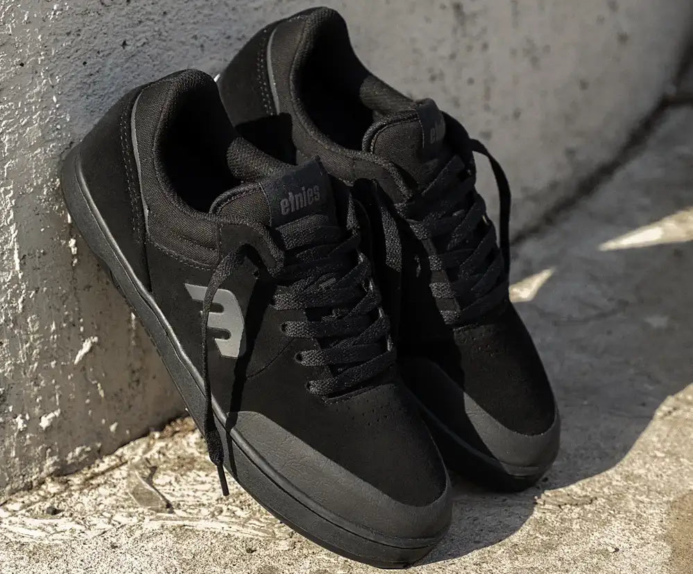 A pair of black low-top sneakers with black laces and a gray side logo, leaning against a textured concrete wall outdoors.