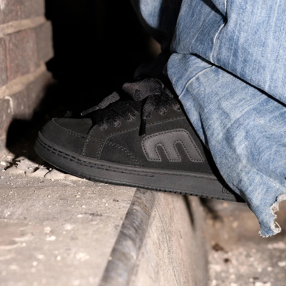 A close-up of a person wearing a black suede sneaker with a tonal gray side logo, paired with frayed blue jeans, sitting on a concrete ledge.