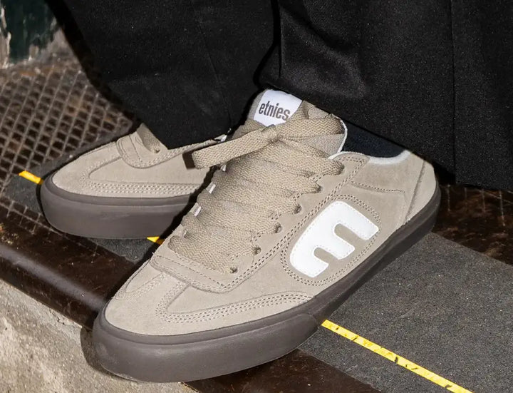 A close-up of a person wearing Etnies low-top sneakers in a taupe suede and dark brown rubber, with black pants visible.