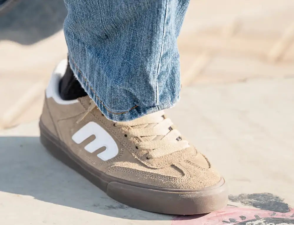 A close-up of a person's foot wearing a tan suede skate shoe with a white logo, paired with blue jeans, standing on a concrete surface.