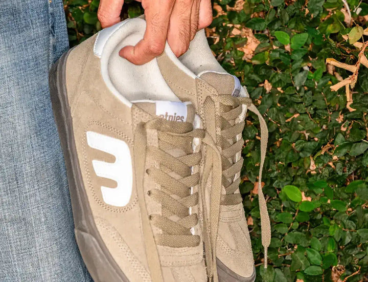 A close-up of a beige suede sneaker held by a hand, with blue jeans visible on the left and green foliage in the background. The shoe has a large white side logo and a white tongue label reading 'etnies'.