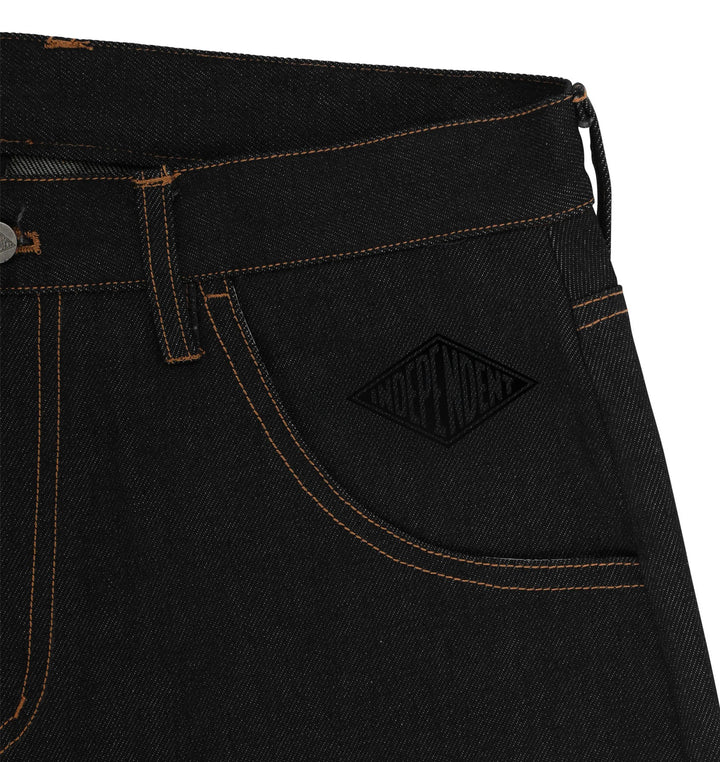 Close-up of black denim jeans showing the waistband, belt loop, front pocket, and a black embroidered diamond-shaped logo patch on the pocket.