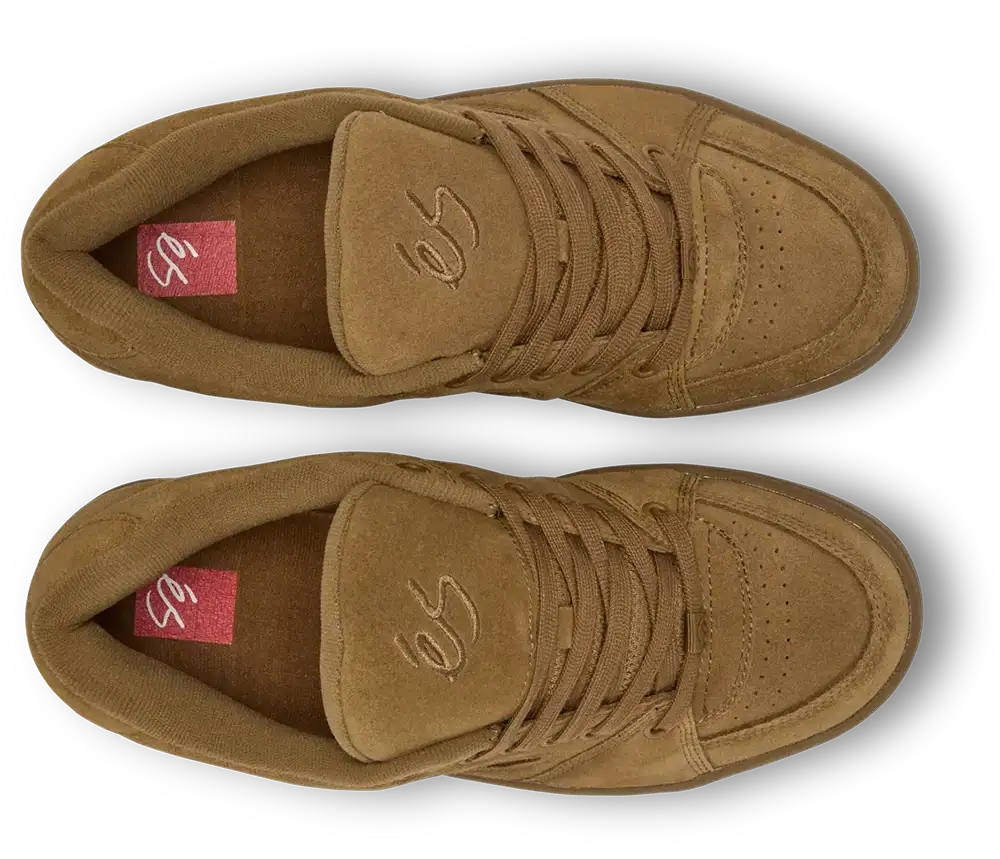 A pair of low-top suede sneakers in a warm tan-brown color, shown from a top-down view on a white background with a soft drop shadow. The shoes feature matching brown laces, a stitched logo on the tongue, and a red insole label.