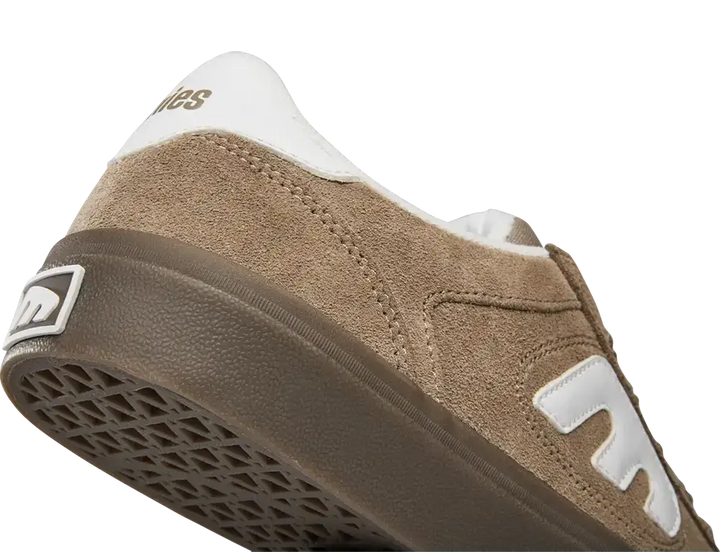 A close-up shot of a New Balance sneaker showing the side profile, featuring a brown suede upper, a white heel tab with gold branding, a white side logo, and a brown rubber sole.