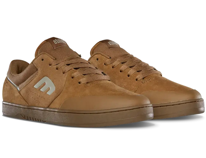 A pair of low-top skate shoes in a warm brown suede and leather mix, featuring a metallic silver 'm' logo on the side, brown laces, and a matching brown rubber sole, shown against a plain white background.