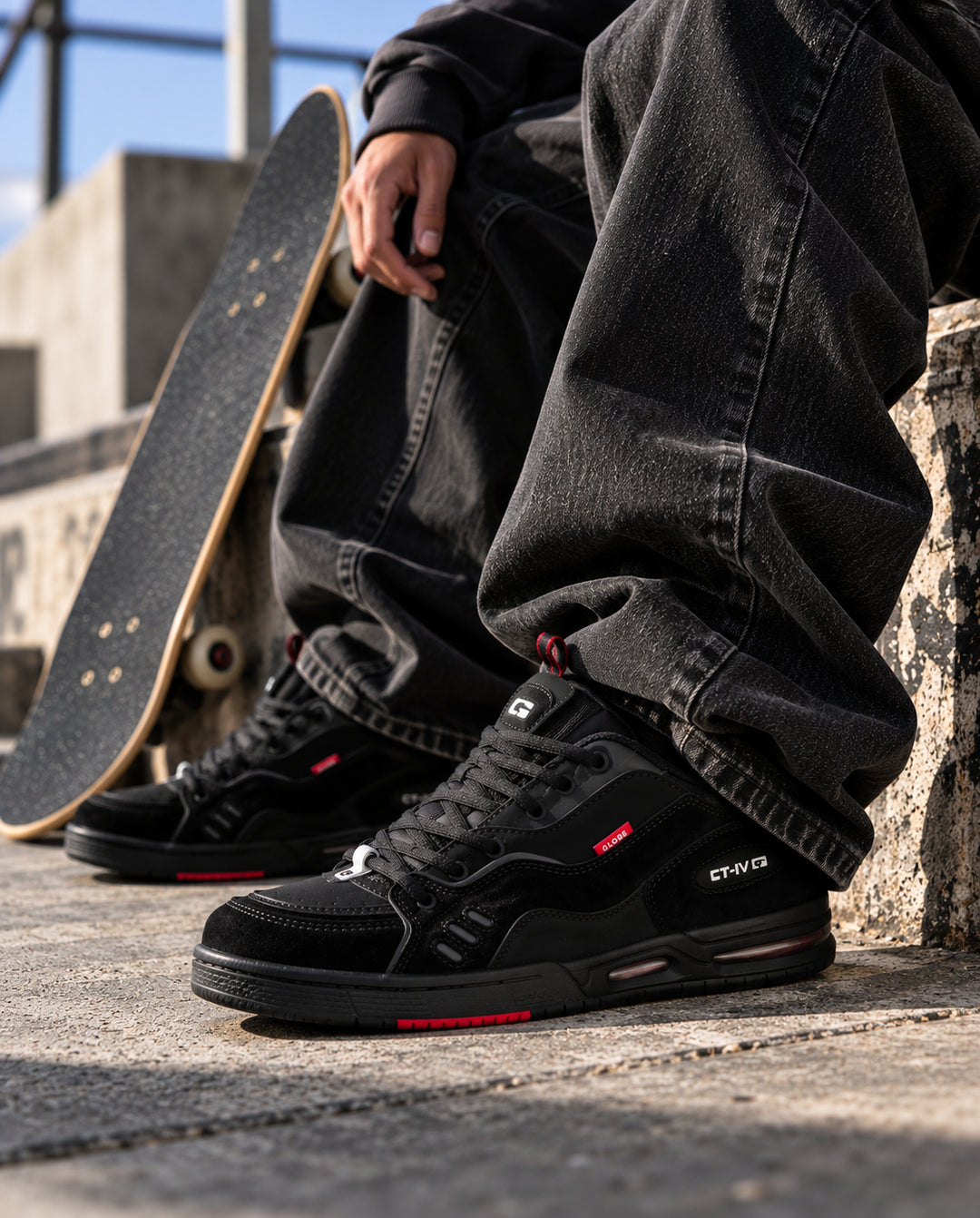 A person wearing black Globe sneakers with red accents and black jeans sits outdoors on a concrete ledge next to a skateboard. The brand name 'CT-IV' is visible on the side of the shoe.