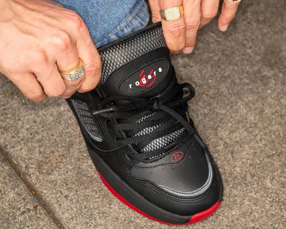 Person wearing a black high-top sneaker with red sole on a sidewalk.