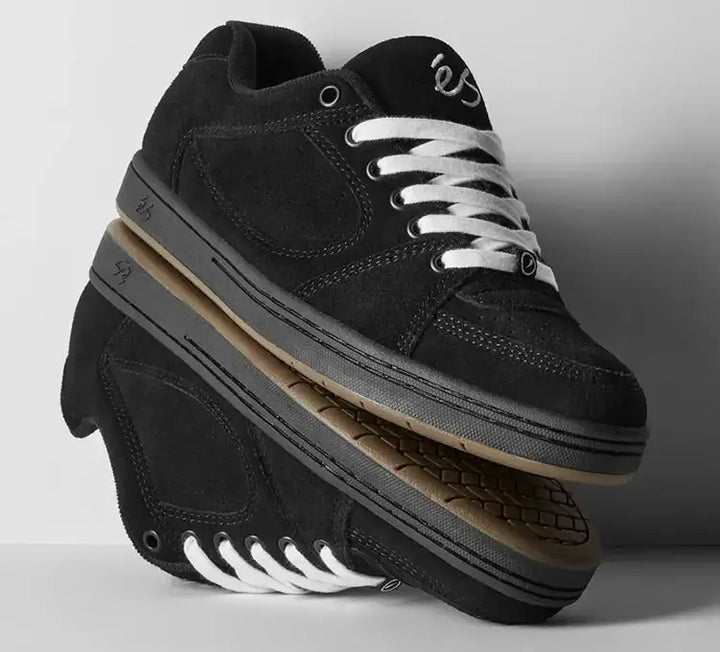Black suede skate shoes with white laces, stacked with one shoe flipped over to show the sole, on a light gray background.