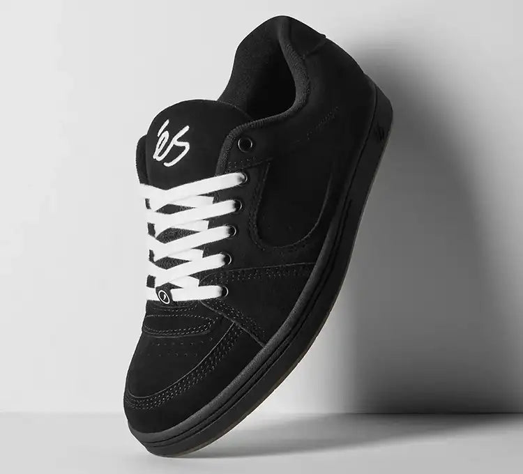 A black low-top sneaker with white laces and a white stylized logo on the tongue, shown floating against a light gray background.