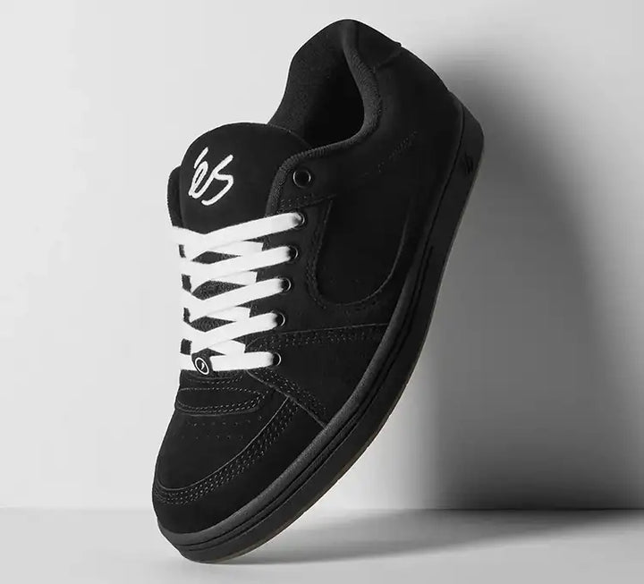 A black low-top sneaker with white laces and a white stylized logo on the tongue, shown floating against a light gray background.