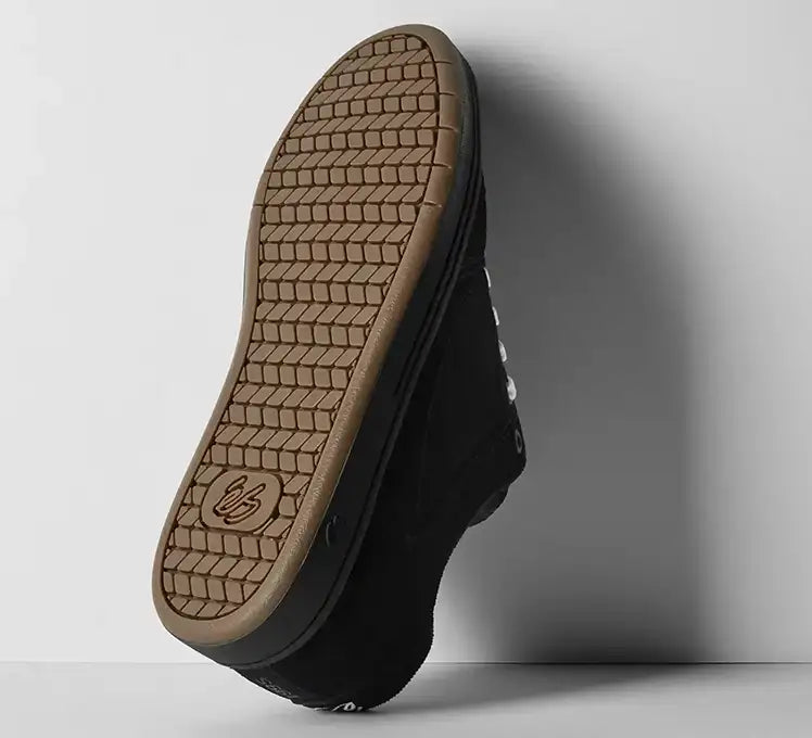 A black low-top sneaker shown from the sole view, leaning against a light gray wall. The tan rubber outsole features a prominent tread pattern and a debossed logo near the heel.