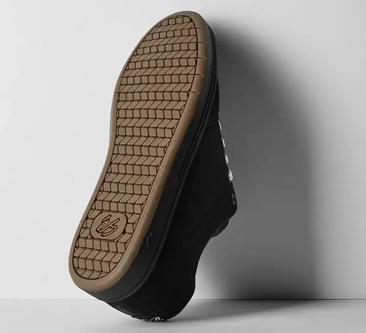 A black low-top sneaker shown from the sole view, leaning against a light gray wall. The tan rubber outsole features a prominent tread pattern and a debossed logo near the heel.