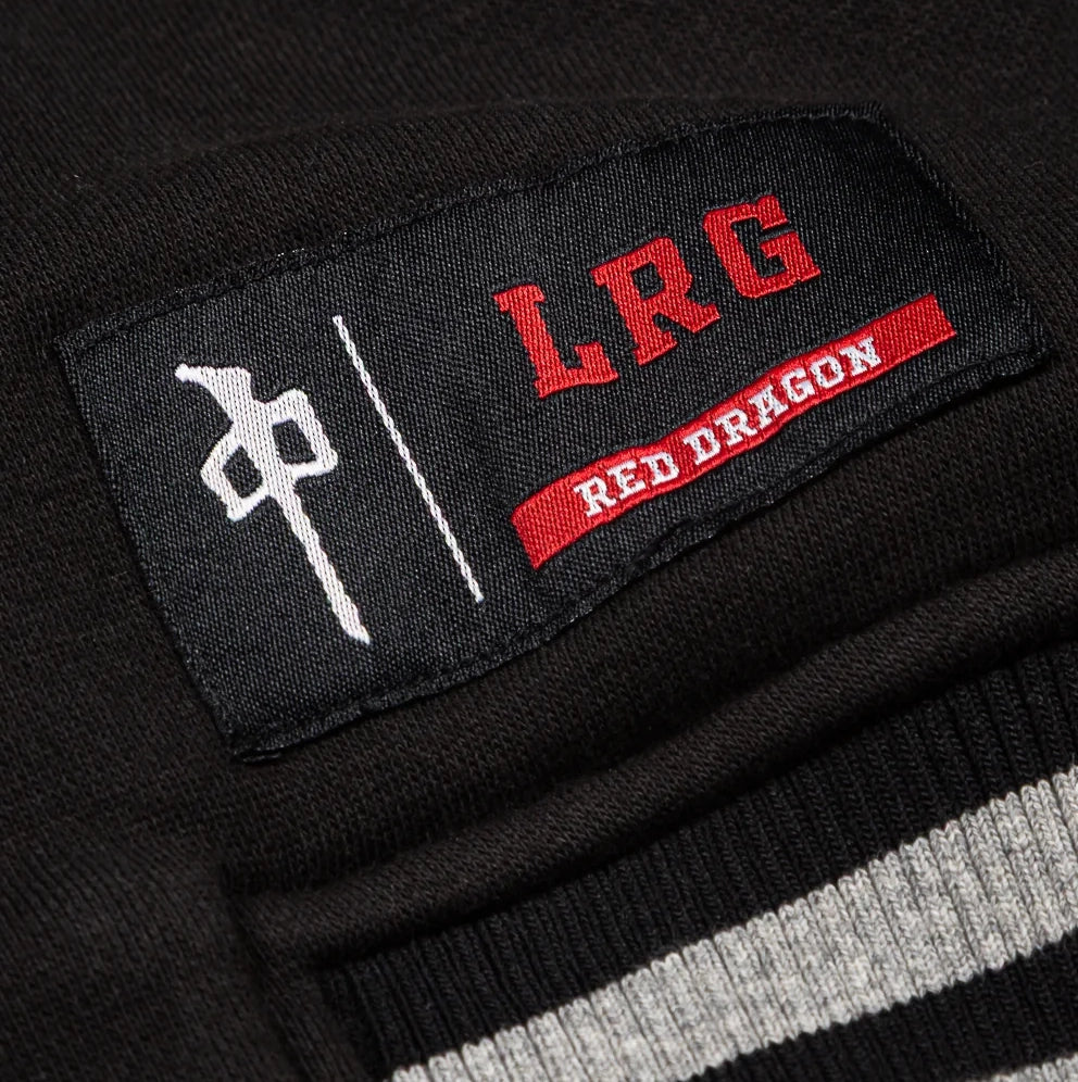 Close-up of a black knit garment patch with red embroidered 'LRG' lettering, a red 'RED DRAGON' banner, and a white symbol, attached to a black ribbed knit collar with white and gray stripes.