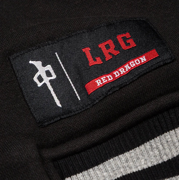 Close-up of a black knit garment patch with red embroidered 'LRG' lettering, a red 'RED DRAGON' banner, and a white symbol, attached to a black ribbed knit collar with white and gray stripes.